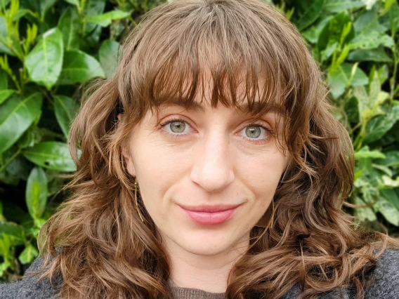 A close-up outdoor portrait of Thea Van Gorp smiling softly in front of a background of lush green foliage. She has wavy, shoulder-length light brown hair with bangs, green eyes, and is wearing a brown top and dark gray coat. The lighting is soft and natural, highlighting her features.