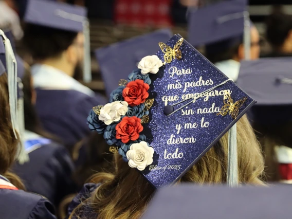 A close-up of a graduation cap decorated with red, white, and navy blue paper flowers, gold butterflies, and glittery blue fabric. In white script, the cap reads in Spanish: ‘Para mis padres que empezaron sin nada y me lo dieron todo’ followed by ‘Class of 2025.’