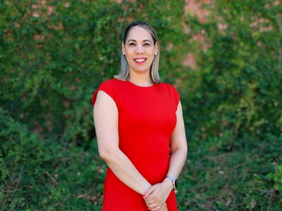Portrait of Maritza Marquez standing outdoors in front of a lush green ivy-covered wall. She is wearing a bright red dress, smiling confidently with hands clasped in front of her. Her straight, shoulder-length hair is styled with blonde highlights.