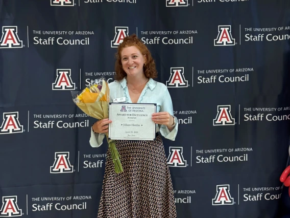 Jillian Hanlin stands smiling in front of a University of Arizona Staff Council backdrop decorated with red, white, and blue balloons, including a large red star balloon. She holds a bouquet of yellow flowers and a certificate titled 'Award for Excellence in Human Resources' dated April 25, 2025.