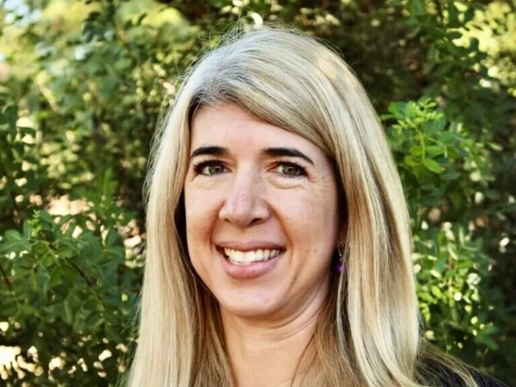 A woman with long, straight, blonde hair smiling at the camera. She is wearing a black top and delicate jewelry, standing outdoors with leafy green foliage in the background