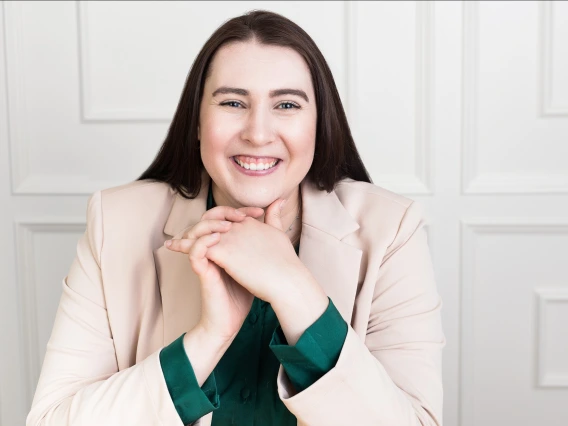 A professional headshot of a smiling individual with long dark brown hair, wearing a light beige blazer over a dark green blouse. They are sitting in front of a white paneled wall with hands clasped and resting under their chin in a friendly, confident pose.