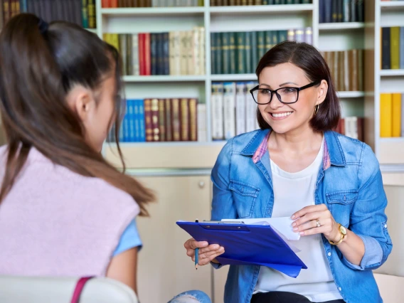 A smiling school counselor wearing glasses and a denim jacket holds a clipboard while talking with a young student. The student, with long brown hair tied in a ponytail, is seated across from the counselor in a room lined with colorful bookshelves. 