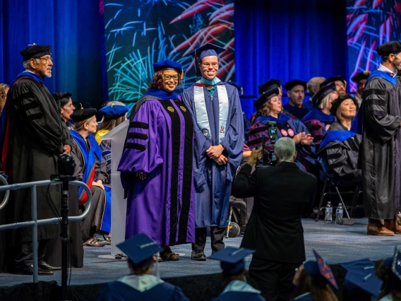 University of Arizona graduation ceremony showing a graduate in a blue cap and gown posing with a faculty member in a purple academic robe, with other faculty and graduates seated on stage in the background and colorful fireworks displayed on screens.