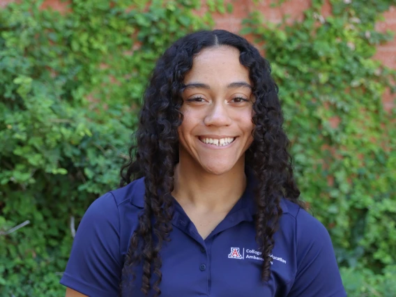 Person with long curly dark hair smiling, wearing a navy blue College of Education Ambassadors polo shirt, sitting in a wheelchair with green vines in the background.
