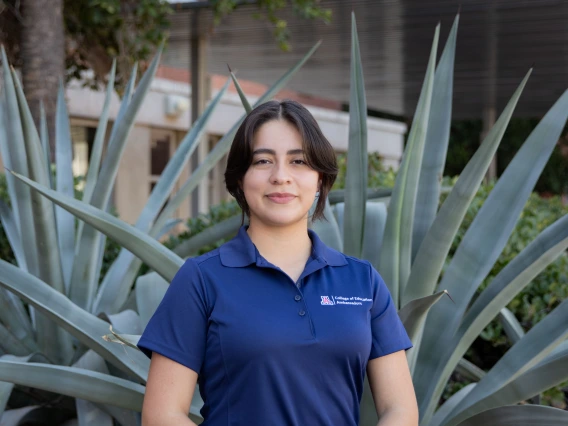 Person with short dark hair smiling, wearing a navy blue College of Education Ambassadors polo shirt, standing in front of large desert plants.