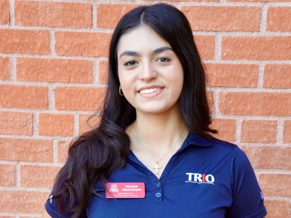 Person with long dark hair smiling, wearing a navy blue polo shirt with a red name tag and TRIO logo, standing against a brick wall.