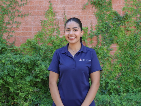 Person with dark hair pulled back smiling, wearing a navy blue College of Education Ambassadors polo shirt, standing in front of a brick wall with green vines.