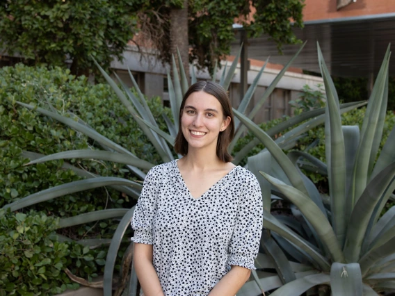 Person with short brown hair smiling, wearing a white blouse with a black pattern, standing in front of large green desert plants