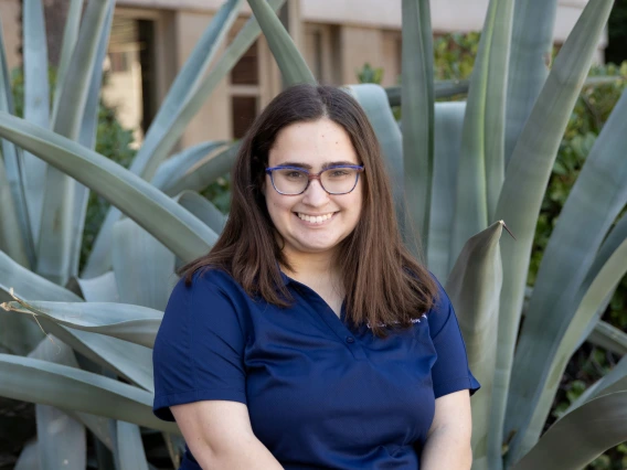 Person with straight brown hair and glasses smiling, wearing a navy blue polo shirt, standing in front of large green desert plants.