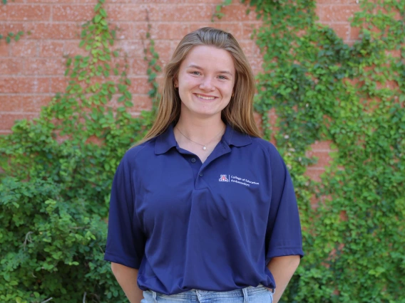 Person with light brown hair smiling, wearing a navy blue College of Education Ambassadors polo shirt, standing in front of a brick wall with green vines.