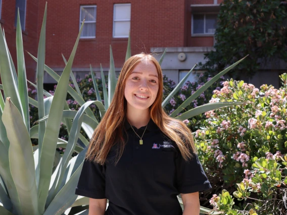 Person with long red hair smiling, wearing a black polo shirt with the College of Education logo, standing outside near large desert plants and pink flowers.