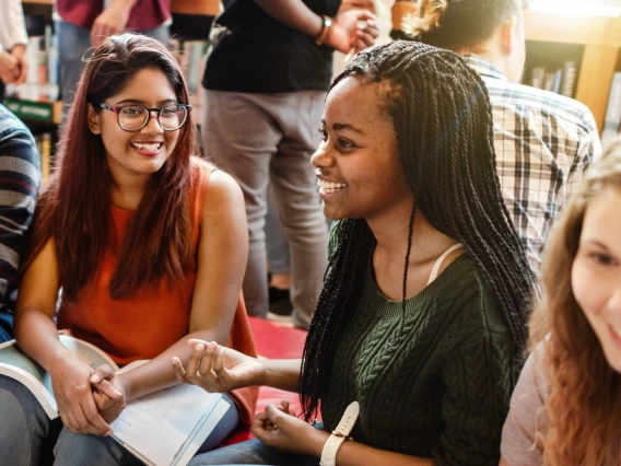 Group of students sitting together in a library, smiling and talking while holding books, conveying connection and shared learning.