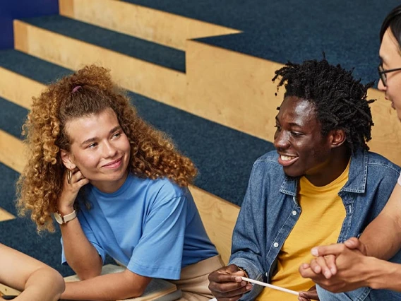 Four diverse students sitting on indoor steps, smiling and talking together with notebooks and headphones, representing collaboration and community in an academic setting.