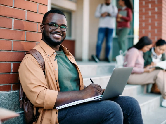 Student smiling and sitting on building steps with a laptop open on their lap.