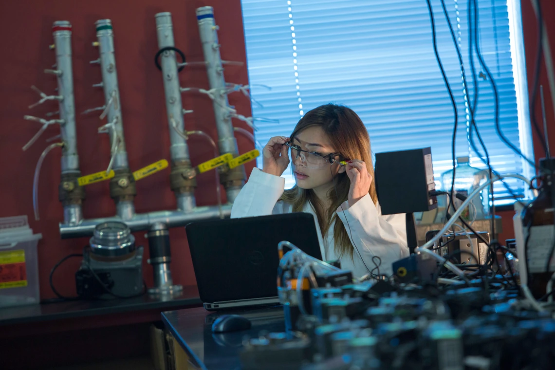 young woman sitting in a lab environment in a lab coat looking at laptop