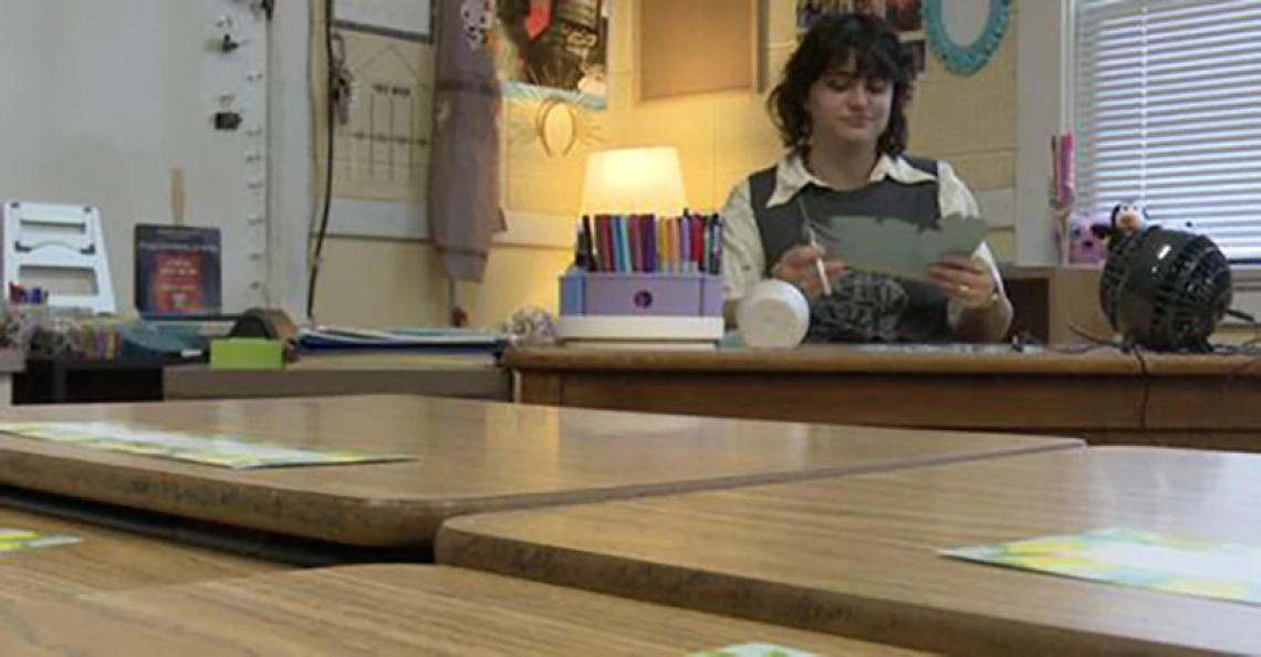 inez mcclain sitting at a teachers desk in a classroom with a lamp lit behind her