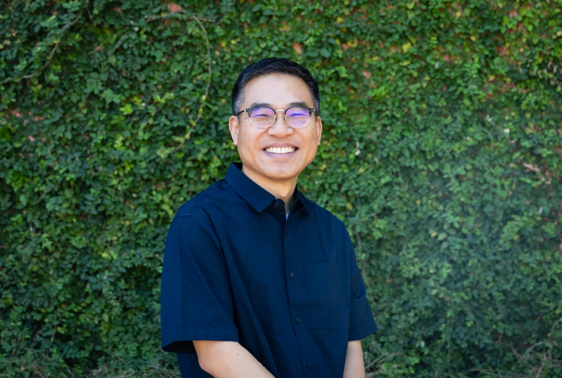 ahn headshot, wearing a blue shirt, eyeglasses, greenery in the background