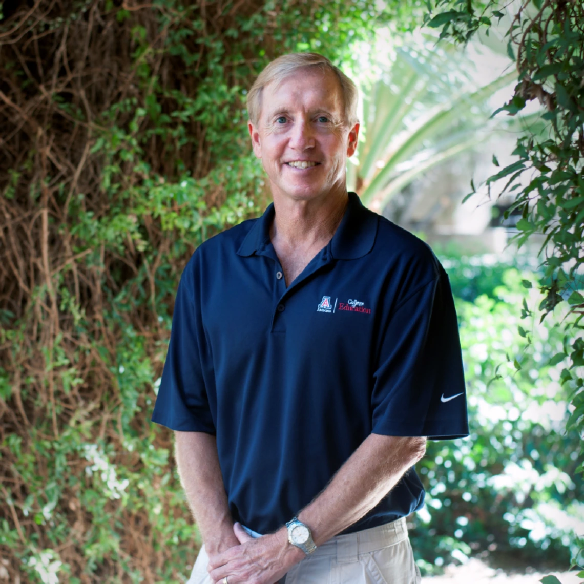 mark jenks headshot, wearing a dark blue UA polo, standing outside with greenery in the background