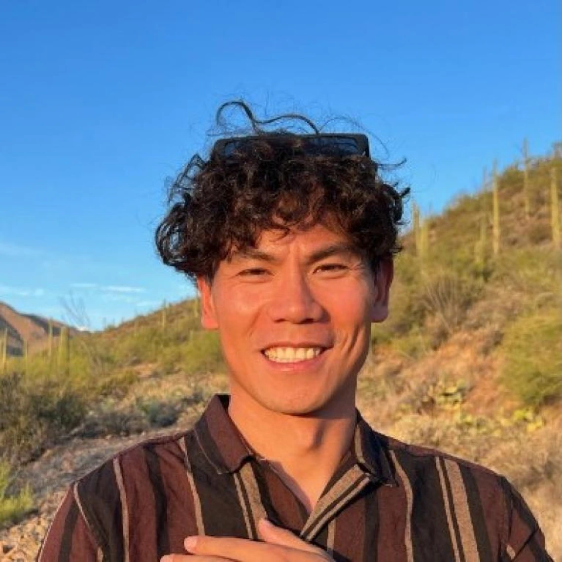 xingxing wu headshot, standing outside under a sunny sky, curly black hair, wearing a striped shirt