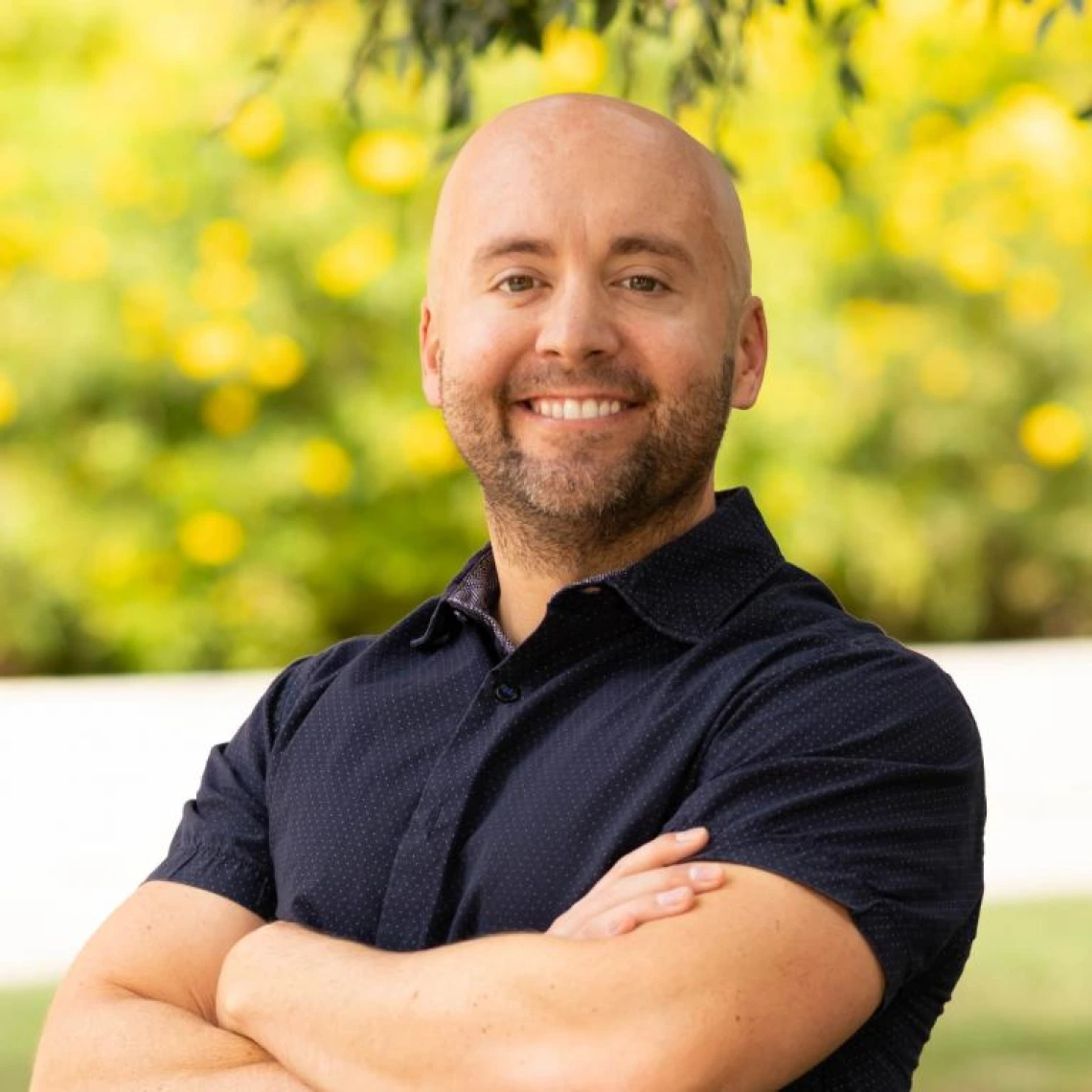 matt ostermeyer headshot, standing outside under a sunny sky, wearing a black polo, arms crossed