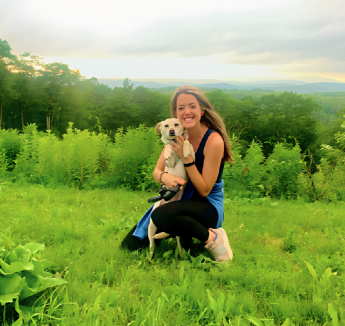 Alison Irwin kneeling in a grassy field, holding a small dog, sunset in the distance