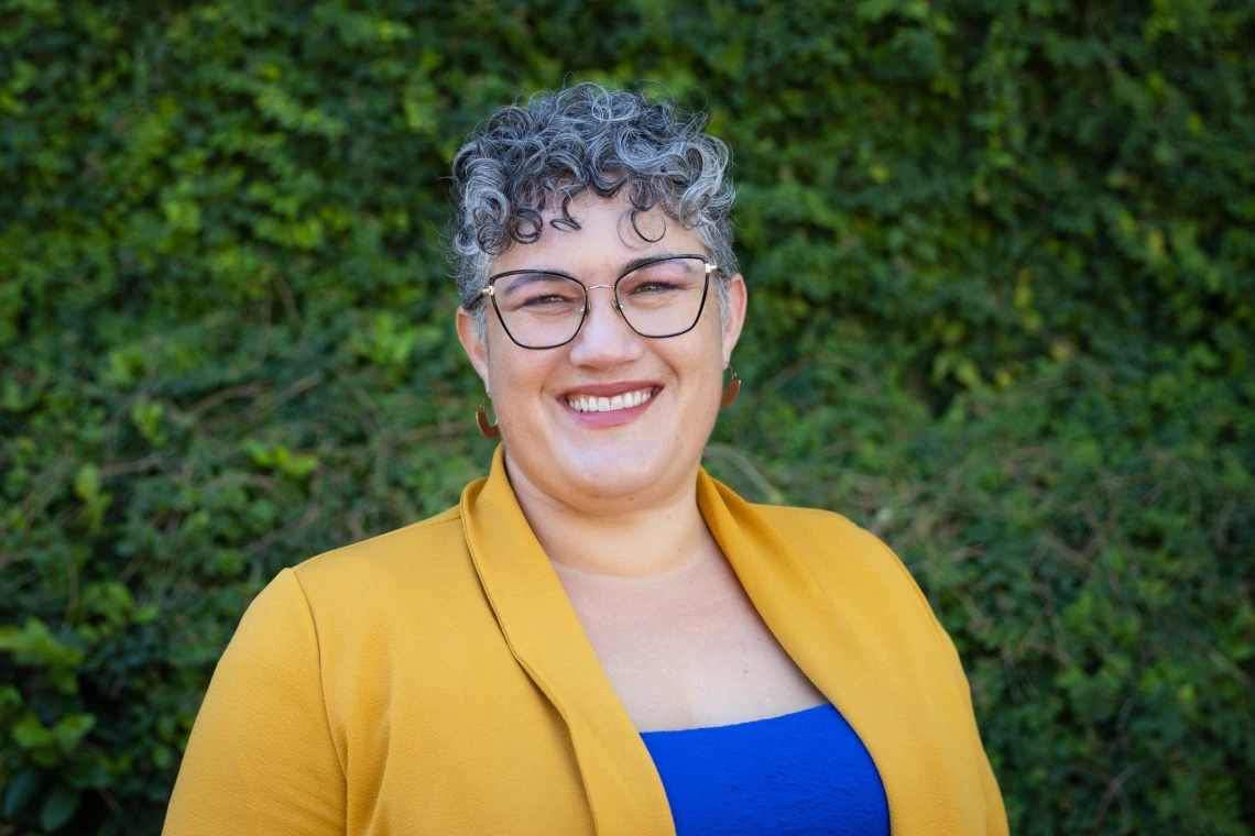 heather wearing yellow jacket, blue blouse, glasses and salt and pepper hair, greenery in background