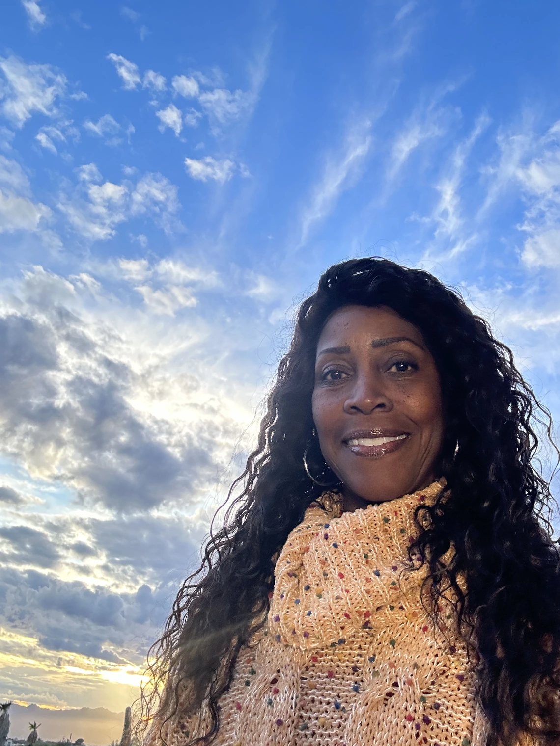 A woman with long, curly hair wearing a beige knitted sweater with colorful speckles, smiling against a scenic backdrop of a blue sky with wispy clouds and a golden sunset.