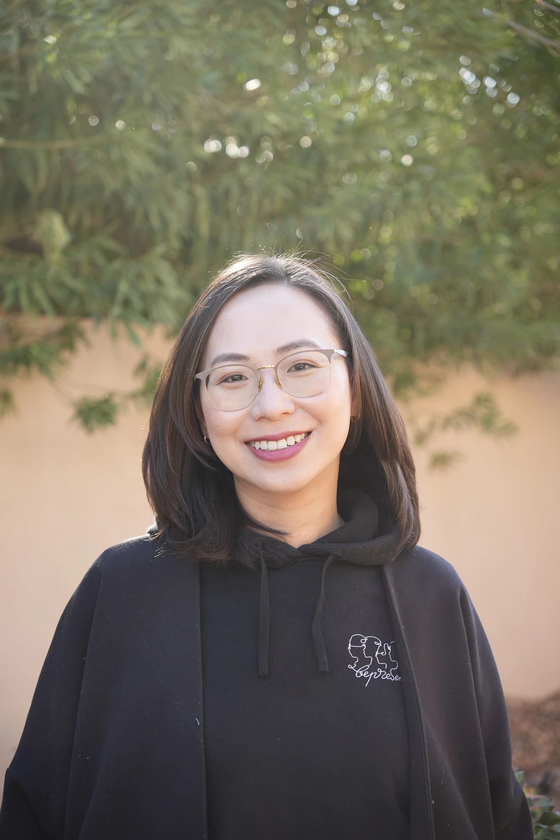 A woman with shoulder-length dark hair and glasses, smiling while wearing a black hoodie with an embroidered design, standing outdoors with green foliage in the background.