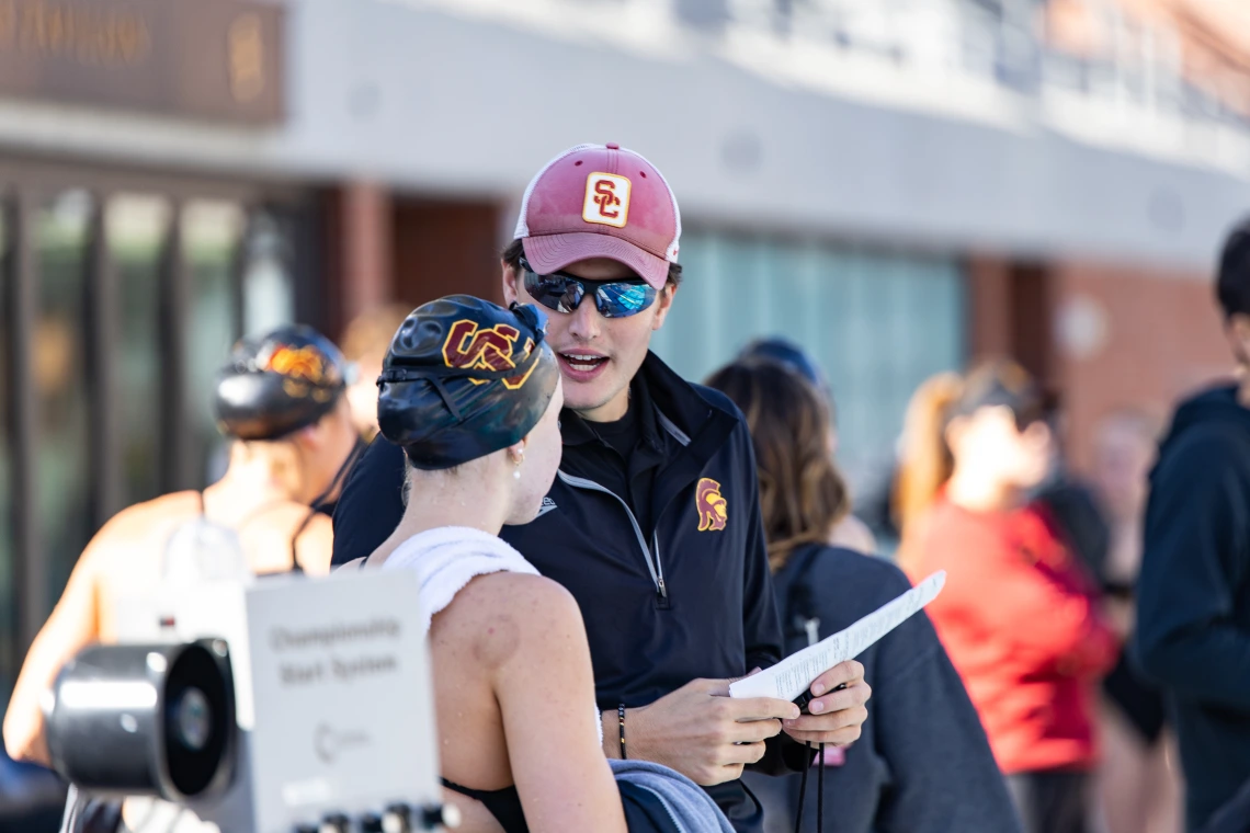 A swim coach wearing a red USC cap, sunglasses, and a black jacket with the USC Trojans logo speaks with a swimmer wearing a black swim cap with the USC logo and a white towel draped over their shoulders. The coach is holding a piece of paper and appears to be giving instructions. Other swimmers and team members are visible in the background at the poolside.