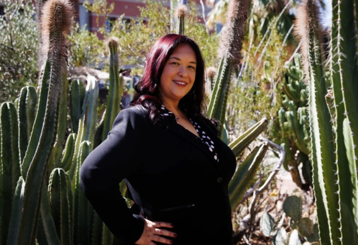 Portrait of Dr. Regina Deil-Amen standing confidently with one hand on her hip in front of tall cacti. She is wearing a black blazer over a patterned blouse and has dark hair with red highlights. The background features a desert landscape with greenery and a partially visible building.