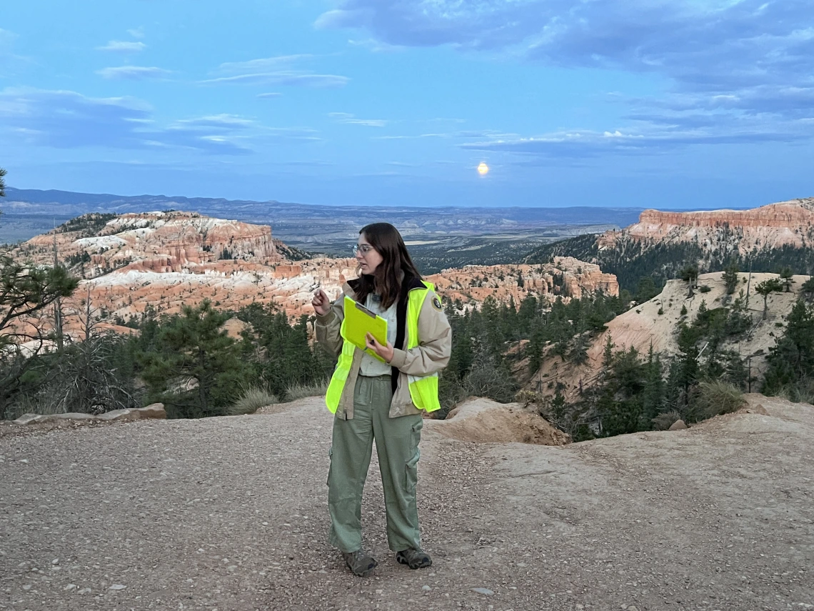 A woman wearing a beige ranger jacket with a patch on the sleeve, a neon yellow safety vest, and green cargo pants stands on a dirt overlook at a national park. She holds a yellow notepad and gestures as if explaining something. The background features a stunning view of red rock formations, green trees, and a full moon rising in the evening sky.