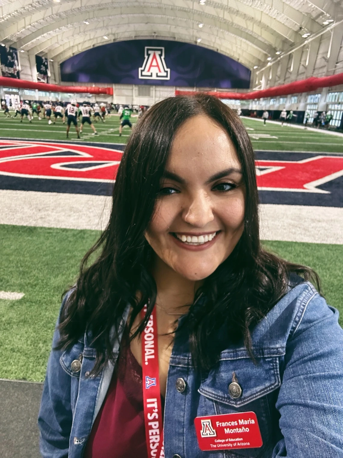 Frances María Montaño smiling on an indoor football field at the University of Arizona, with players practicing in the background. She is wearing a red UA lanyard and a name badge from the College of Education.