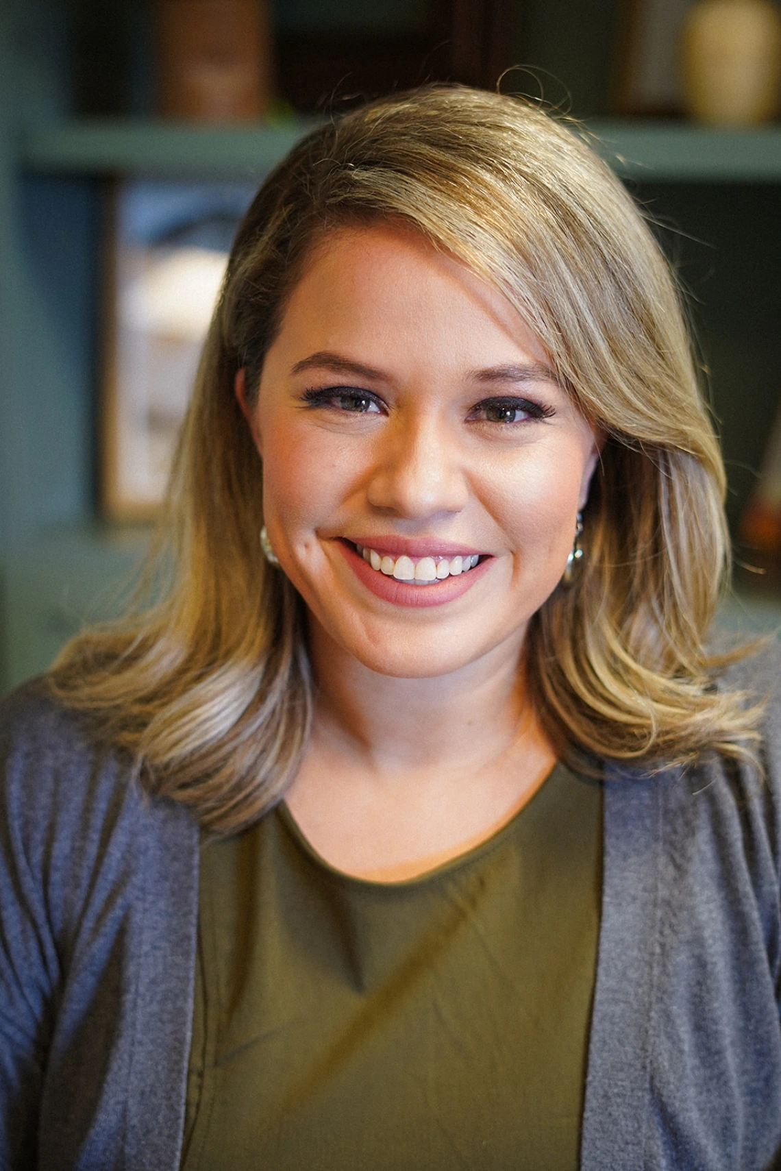 A warm, professional portrait of Laura Torres smiling directly at the camera. She has shoulder-length blonde hair styled to the side and is wearing a dark olive green top with a gray cardigan