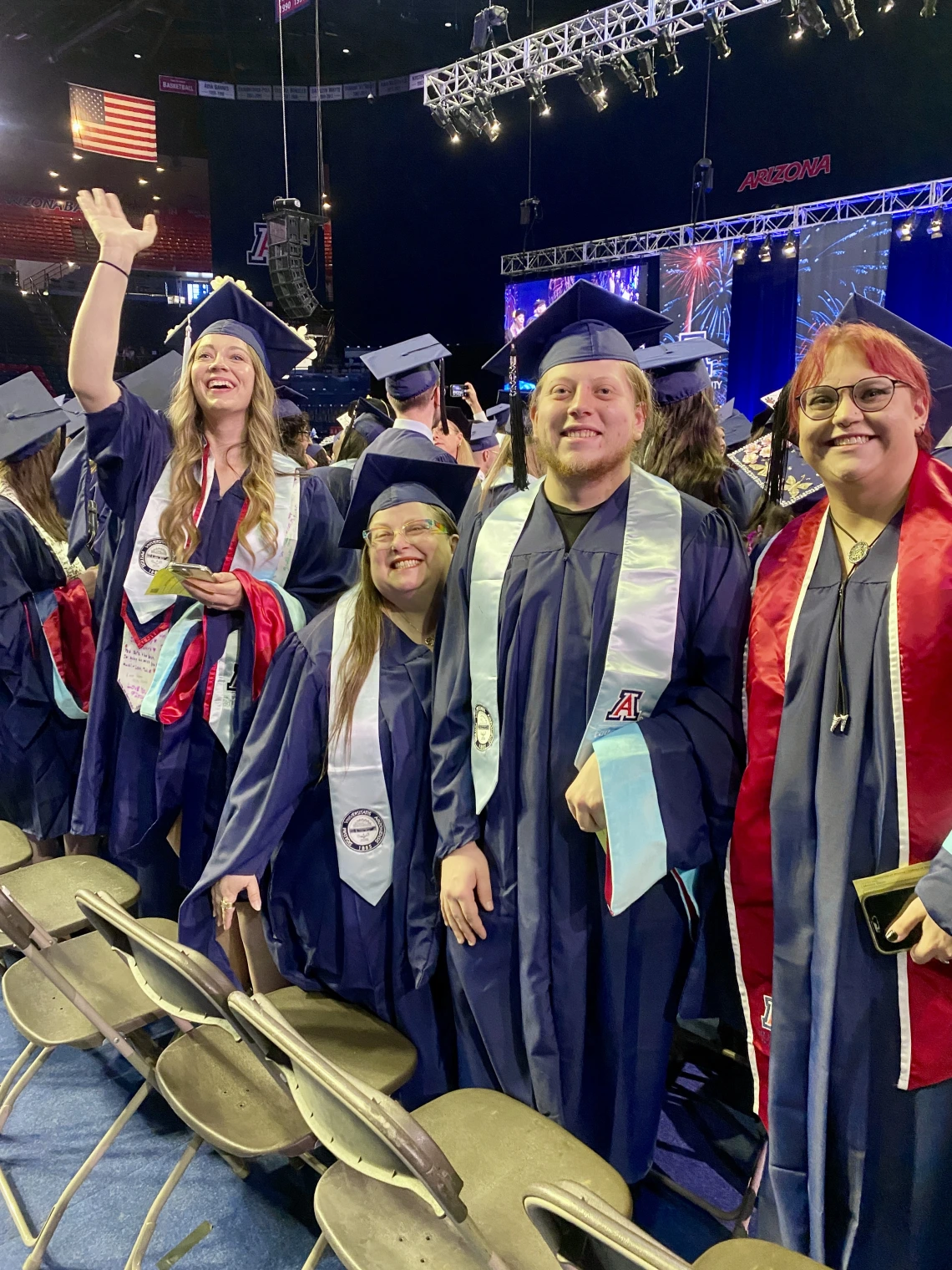 A group of smiling University of Arizona graduates in navy blue caps and gowns stand together during a commencement ceremony. One graduate waves excitedly while others smile at the camera.