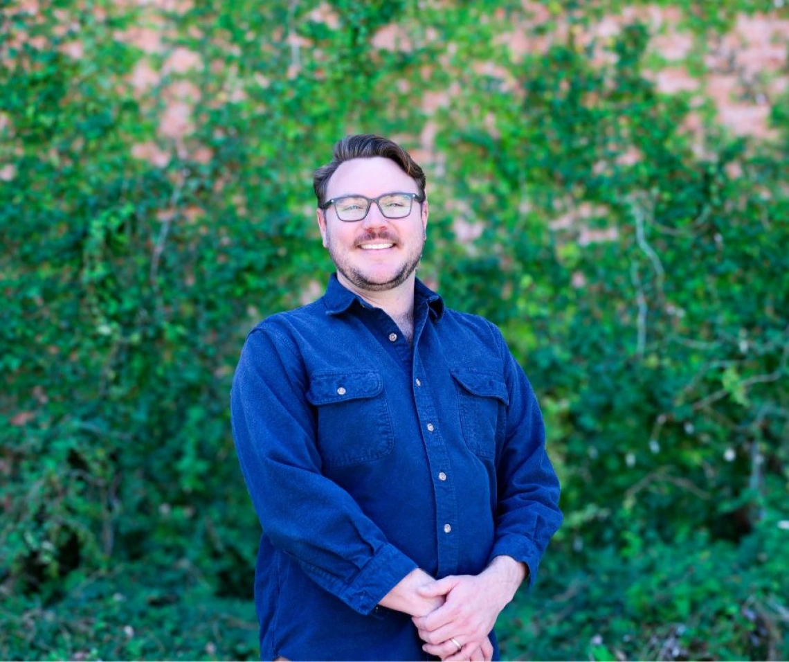 Portrait of Kyle Fisher smiling and standing in front of a vibrant ivy-covered wall. He is wearing glasses and a dark blue button-up shirt, with his hands loosely clasped in front. His hair is styled neatly, and he has a short beard.