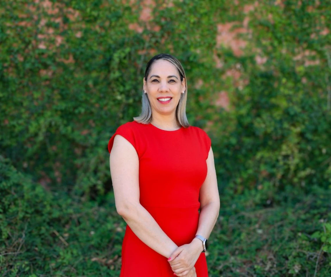 Portrait of Maritza Marquez standing outdoors in front of a lush green ivy-covered wall. She is wearing a bright red dress, smiling confidently with hands clasped in front of her. Her straight, shoulder-length hair is styled with blonde highlights.