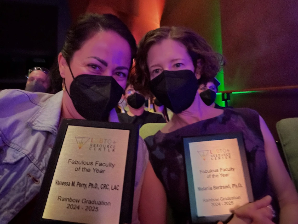 Vanessa Perry and Melanie Bertrand sit side by side in an auditorium, each holding a plaque awarded by the LGBTQ+ Resource Center. Both are wearing black face masks and smiling with their eyes.