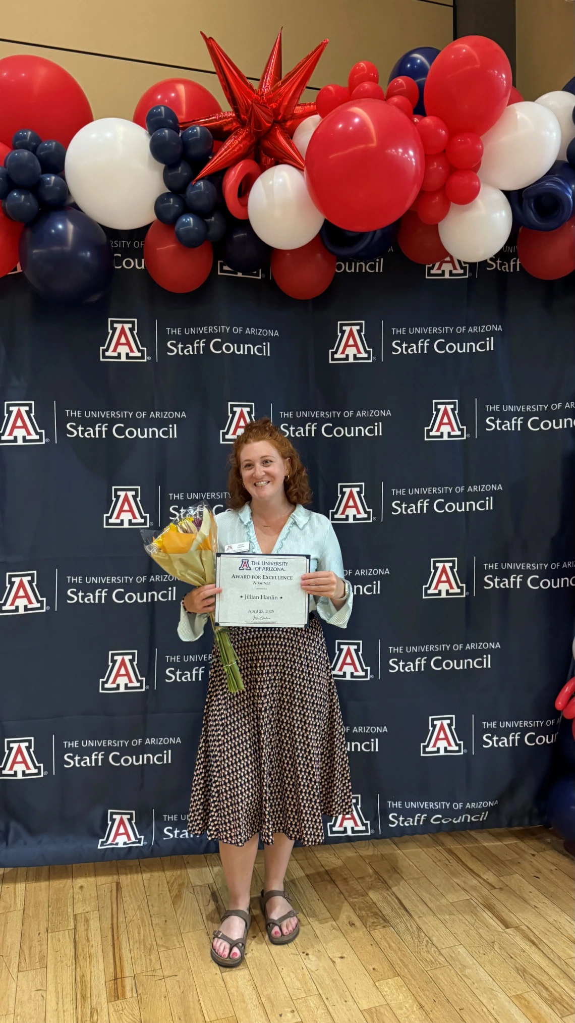 Jillian Hanlin stands smiling in front of a University of Arizona Staff Council backdrop decorated with red, white, and blue balloons, including a large red star balloon. She holds a bouquet of yellow flowers and a certificate titled 'Award for Excellence in Human Resources' dated April 25, 2025.