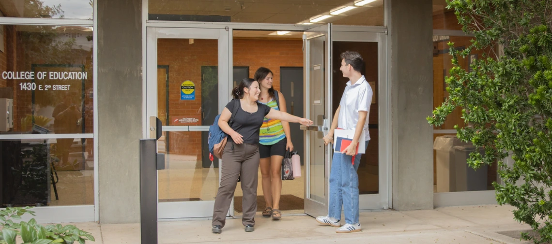 Three college students stand outside the entrance of the College of Education at 1430 E. 2nd Street. One male student holds the door open while smiling and speaking with two female students, one of whom is pointing inside. All three appear to be engaged and friendly, with backpacks and books in hand.