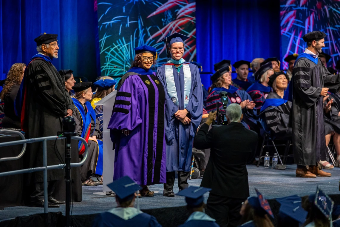 University of Arizona graduation ceremony showing a graduate in a blue cap and gown posing with a faculty member in a purple academic robe, with other faculty and graduates seated on stage in the background and colorful fireworks displayed on screens.