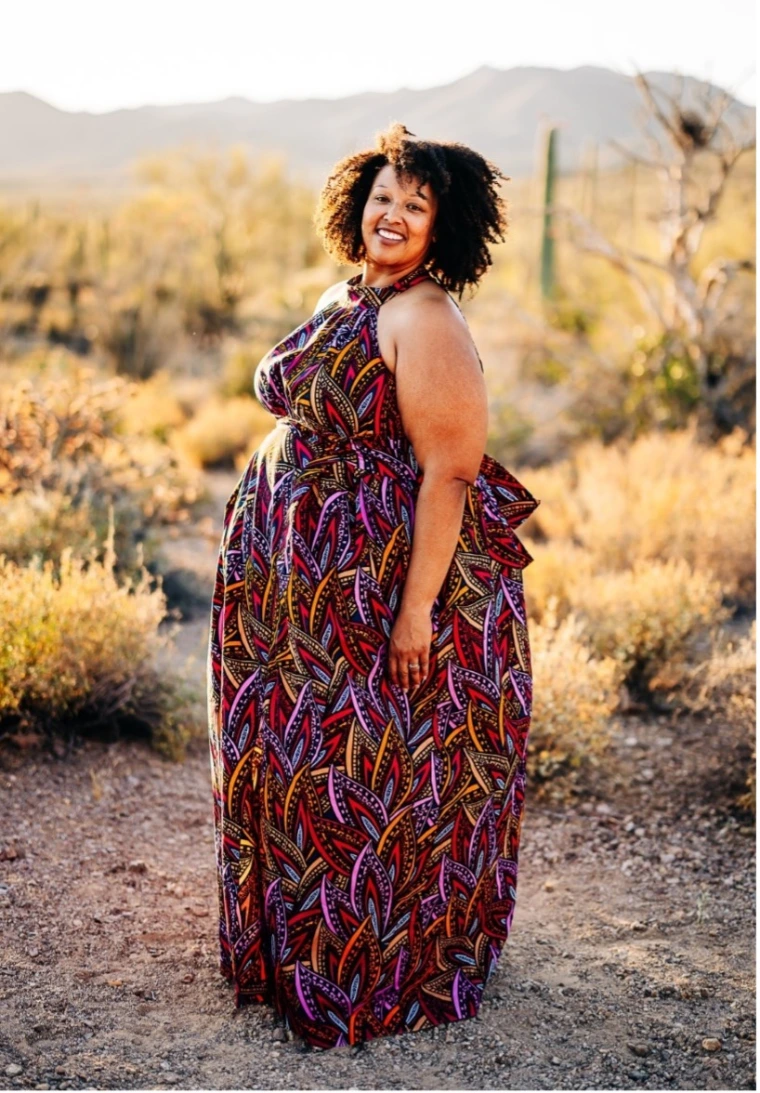 A smiling woman with curly black hair stands in a desert landscape during golden hour. She is wearing a colorful, patterned maxi dress with a high neckline. The background features cacti, desert shrubs, and distant mountains bathed in warm sunlight.