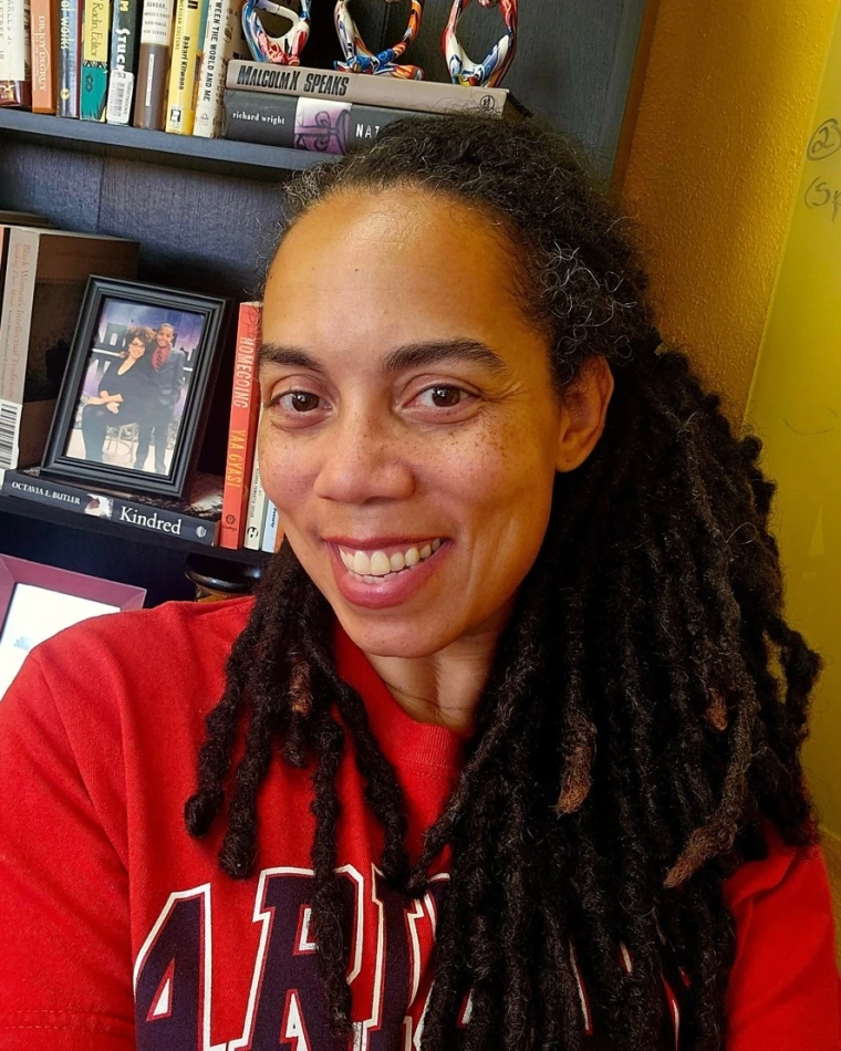 A smiling woman with long, dark dreadlocks wearing a red University of Arizona shirt takes a selfie in front of a bookshelf filled with books, awards, and a framed photograph