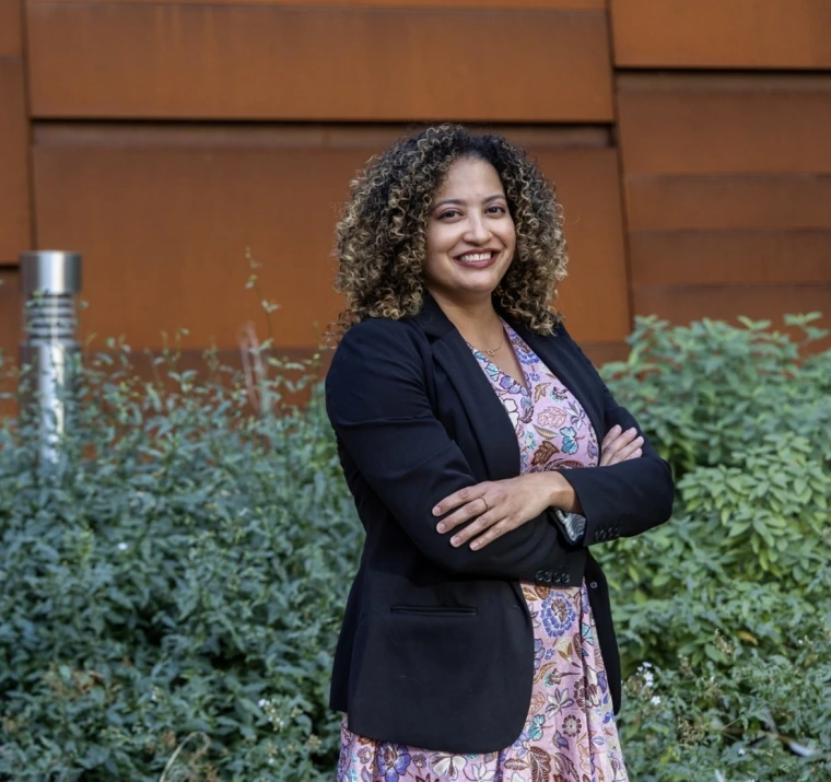 Portrait of Dr. Desiree Vega, a woman with curly brown hair, smiling and posing with arms crossed. She is wearing a black blazer over a floral-patterned dress. The background features green plants and a modern rust-colored metal wall.