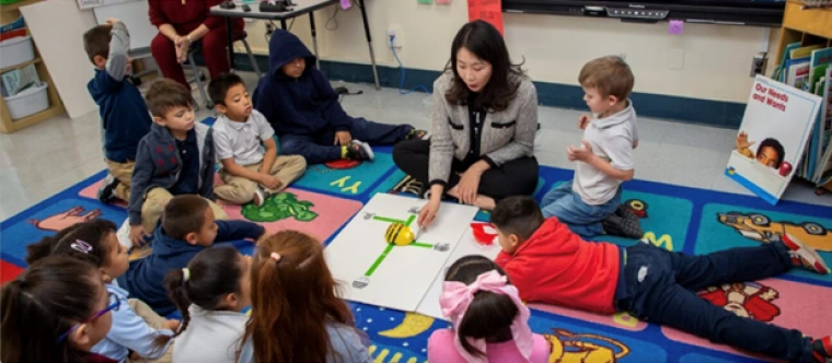 Dr. Sung Eun Jung sits on a colorful classroom rug surrounded by young children as she teaches a hands-on activity using a bee-shaped robot on a directional map. The children are engaged, seated or lying on the rug around her.