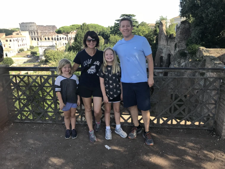 Lia Falco and her family pose together in front of a scenic overlook with the Colosseum and Roman ruins in the background. The family of four—two adults and two young girls—smile at the camera on a sunny day. Lia is wearing a navy Wildcats shirt and black shorts, while her partner wears a light blue shirt and navy shorts. The children are dressed in casual summer outfits. Green trees and ancient stone structures frame the background.