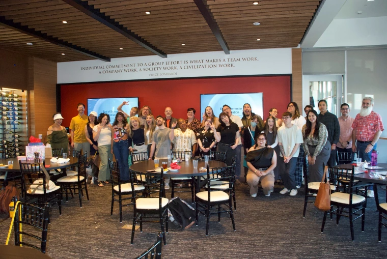 A large group of about 30 people pose playfully for a group photo in a modern event room, many making funny faces or expressive gestures.