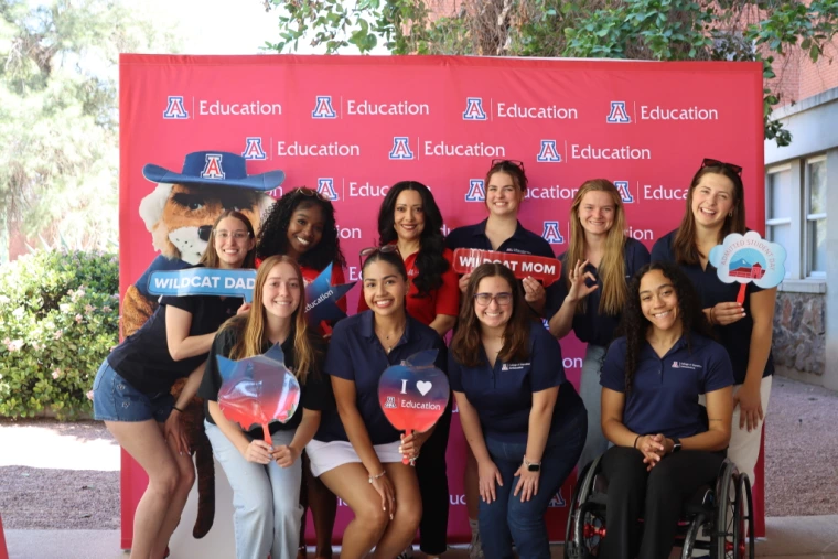 A group of ten smiling University of Arizona College of Education student ambassadors pose in front of a red backdrop that reads ‘Education’ with multiple university logos. They are holding props with phrases like ‘Wildcat Dad,’ ‘Wildcat Mom,’ and ‘I ❤️ Education.’ One student is in a wheelchair, and several are wearing navy College of Education polo shirts.
