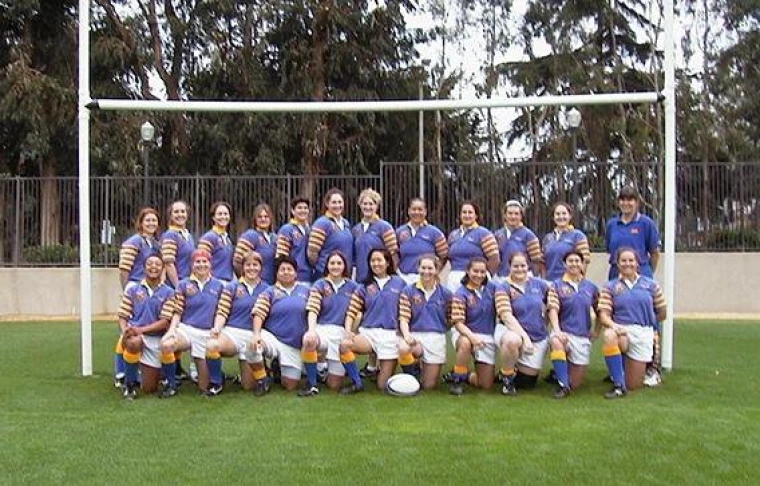 Group photo of the UCLA Women’s Rugby Football Club posed under a goalpost on a grass field. The team members wear matching purple jerseys with yellow-and-white striped sleeves, white shorts, and yellow socks. Cindy Cruz is among the players, all smiling confidently.