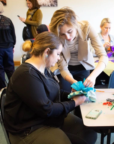 woman sitting at a table working on a project while another leans over standing next to her helping, in classroom setting